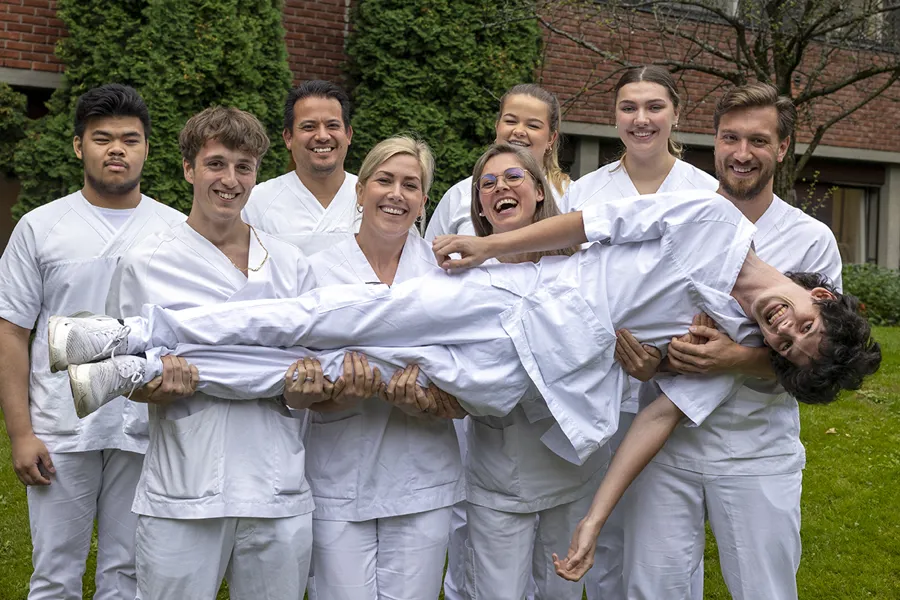 Åtte portører, ledere og lærlinger i hvitt ståri atriet utenfor sykehuset og bærer en portør, som ligger på langs i armene deres