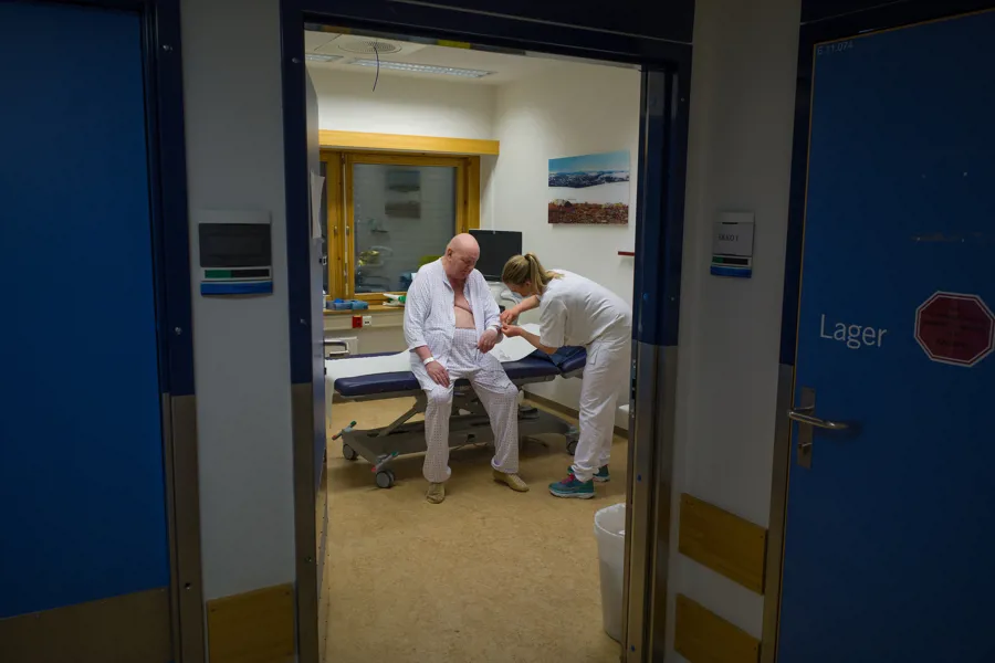 Diakonisykepleier som behandler en pasient på Diakonhjemmet sykehus. Pasienten er en voksen mann som sitter på en sykeseng.