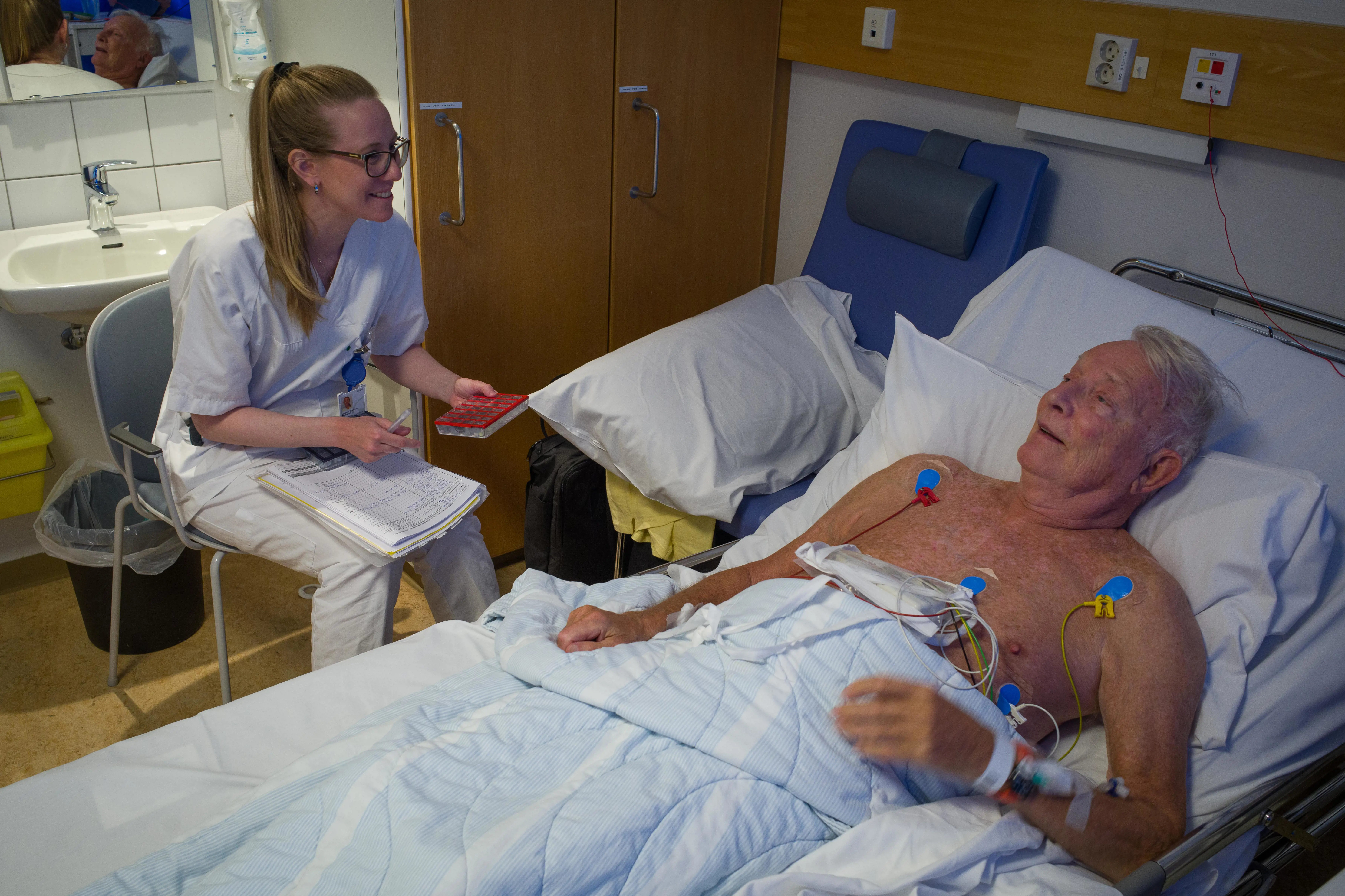 Smilende kvinnelig sykepleier som gir medisiner til en eldre mann. Han ligger i sykeseng.