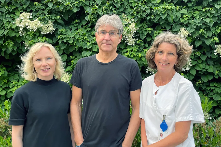 Tre mennesker står foran en beplantet vegg (to damer og en mann).