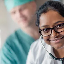 En lege med stetoskop smiler, i bakgrunnen ser man en kirurg.